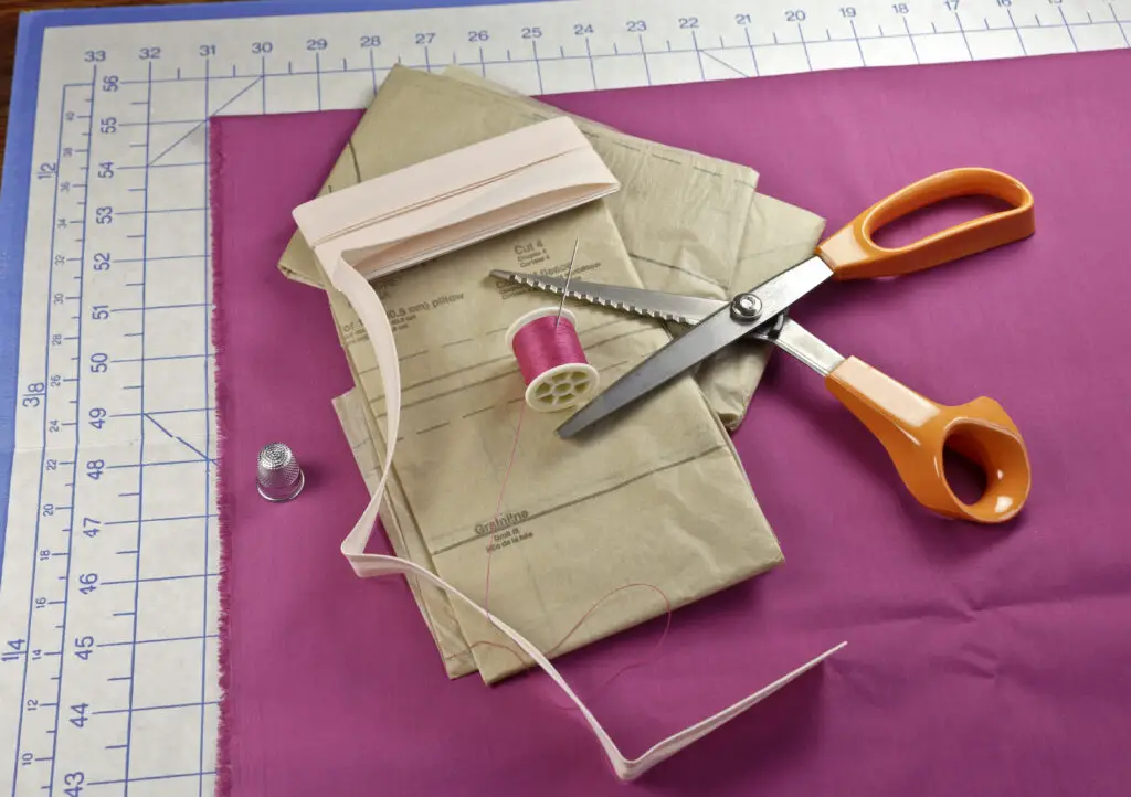 Getting ready to sew by preparing your fabric and pattern. Pink Fabric, Pattern, pinking shears. thimble, thread, needle, cutting board, and bias tape.