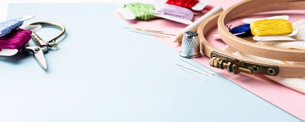 Embroidery accessories that can be used for cross stitching. White fabric, embroidery hoop, colorful threads, scissors and needles. On blue background.